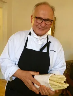 En person i vit kockjacka och svart förkläde håller flera ljusa arbetshandskar i ett rum inomhus..