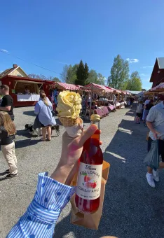 Hand håller en mjukglass och en flaska läsk på en solig utomhusmarknad med stånd och besökare..