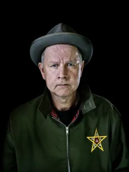 En man med en grå hatt på huvudetiklädd en grön jacka med en gul stjärna på bröstet, svart bakgrund.