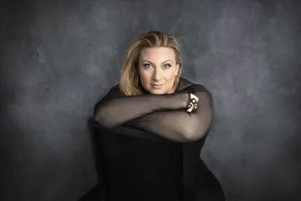 En kvinna med långt hår, iklädd en svart tröja med genomskinliga ärmar, sitter med armarna i kors lutade mot ryggstödet på en stol.