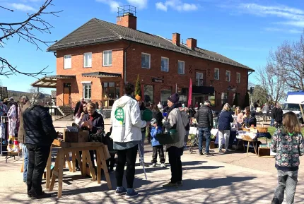 Bord med varor till försäljning, besökare som går runt mellan borden, en röd tegelbyggnad i bakgrunden.