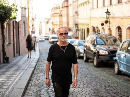 En man med grått hår, skägg och solglasögon iklädd svarta jeans och svart t-shirt, i bakgrunden en kullerstensgata med parkerade bilar och stenhus. 