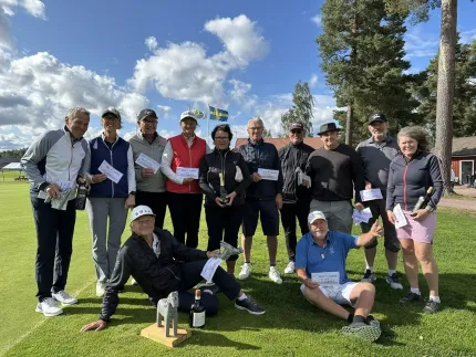 Grupp personer på en golfbana håller diplom och flaskor, med svensk flagga, träd och blå himmel i bakgrunden..