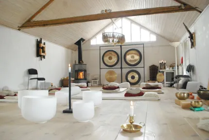 Ljus träningssal med mattor och kuddar på golvet, vita klangskålar i förgrunden och stora gongar vid väggen..