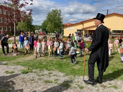 Utomhussamling med barn och vuxna på gräsmatta; en kostymklädd person med hög hatt och käpp står i förgrunden..