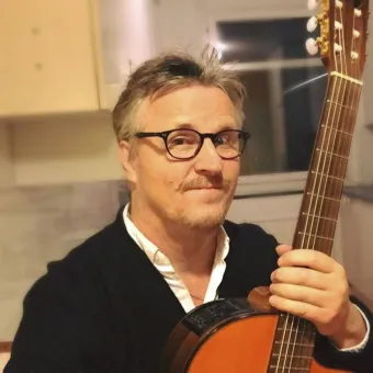 En person i mörk tröja håller en akustisk gitarr inomhus, med köksskåp och fönster i bakgrunden..