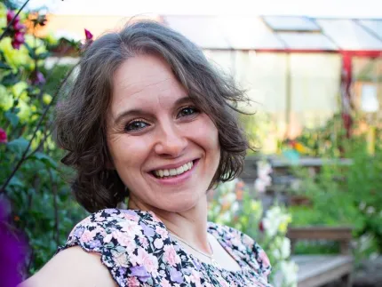 En person i blommig klänning sitter avslappnat på en bänk i en grön trädgård med växthus i bakgrunden..