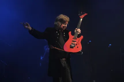 En musiker på en mörk scen håller upp en röd elgitarr medan blå scenbelysning och mikrofonstativ syns..