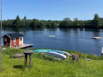 Gräsmatta vid en sjö med rödt båthus på brygga, uppdragna kajaker och skog på andra sidan i sol..