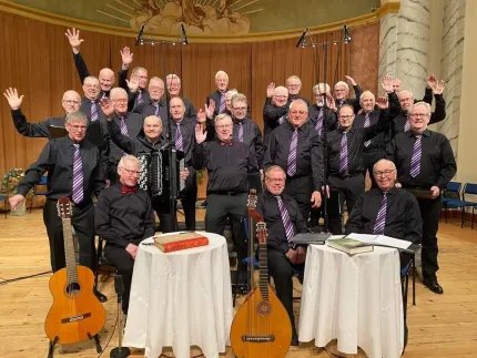 En grupp män i svarta skjortor och svarta byxor och lila slipsar, en gitarr, en luta och två bord med vita dukar  i framkant.