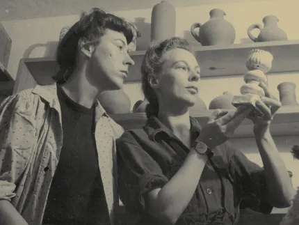 Två personer i en keramikverkstad, en håller upp ett litet keramikföremål; hyllor med krukor i bakgrunden..