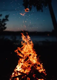 En stor brasa med orange lågor och gnistor som stiger i skymningsljus, med suddiga träd i bakgrunden..