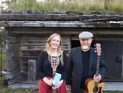 Två personer står framför en gammal timmerstuga och håller akustiska gitarrer; grästak och skog syns i bakgrunden..
