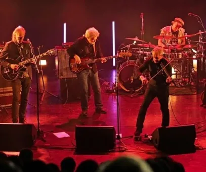 Fyra musiker spelar live på scen med gitarr, bas, violin och trummor i röd belysning framför publik..