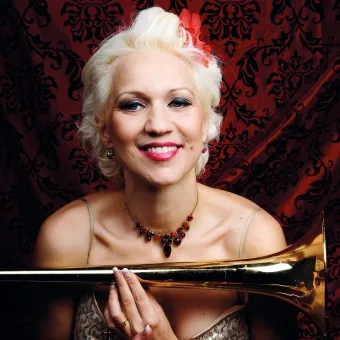 Närbild av en person med blond lockig frisyr och halsband, som håller en trombon framför en röd mönstrad bakgrund..