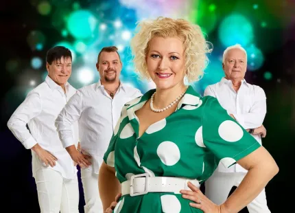 En kvinna med kort ljust hår iklädd en grön klänning med vita prickar, bakom henne tre män iklädda vita skjortor och vita jeans, diskokulor i bakgrunden.