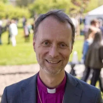 Porträtt av en person i kavaj och lila prästskjorta med vit krage; ansiktet är maskerat, folk i park syns bakom..