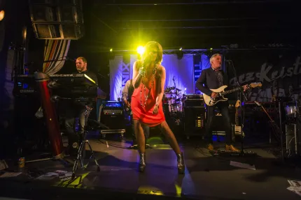 En sångare i röd klänning står i strålkastarljus på scen, med musiker och instrument i bakgrunden..