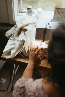 En person syr ljus tyg på en symaskin vid ett fönster, med mycket tyg samlat på bordet i dagsljus..