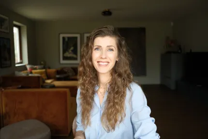 En kvinna med brunt långt lockigt hår, ikädd en blå skjorta, ett vardagsrum i bakgrunden.