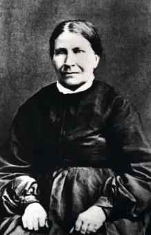 Svartvitt porträtt av en sittande person i mörk klänning med vita manschetter mot en enkel bakgrund..