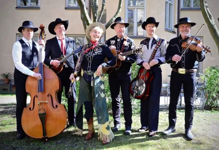 Fem män med cowboyhattar, alla håller i varsitt instrument, i mitten en kvinna i cowboykläder.