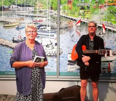 Två personer står framför en målad hamnscen med båtar; den ena håller en bok och den andra en gitarr..