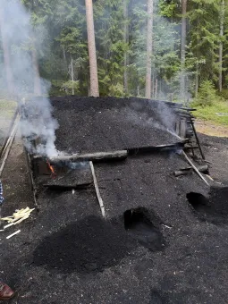 En stor kolmila täckt av svart jord ryker i en barrskog, med trästöd, glöd och små lågor vid kanten..