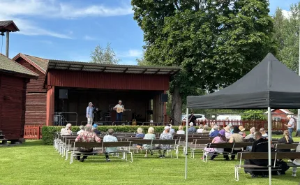 Publik sitter på bänkar framför en röd utomhusscen där två musiker uppträder; stort träd och tält syns..