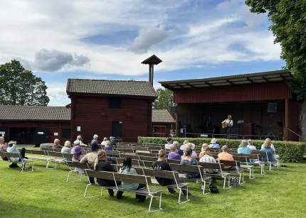 Publik sitter på bänkar på en gräsmatta och lyssnar på en musiker som spelar på en utomhusscen vid röda byggnader..