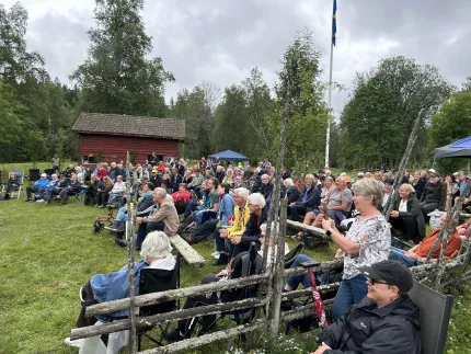 Stor publik sitter på stolar på en gräsmatta vid en röd stuga, med tält, träd och svensk flagga i bakgrunden..