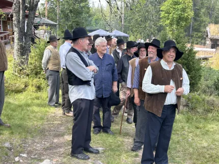 Grupp män i västar och hattar står på en grusväg vid en stuga, omgivna av träd och grönska..