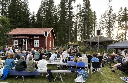 Människor sitter på bänkar och stolar vid en utomhuskonsert nära en röd stuga, omgiven av tallskog..