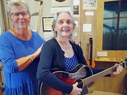 Två personer i blåprickiga klänningar inomhus; en håller en akustisk gitarr medan den andra står bakom..