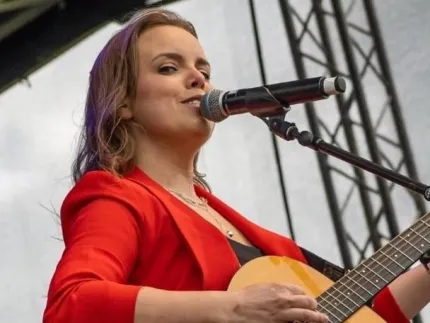 Person i röd kavaj står på en scen och spelar akustisk gitarr vid en mikrofon, med scenrigg i bakgrunden..