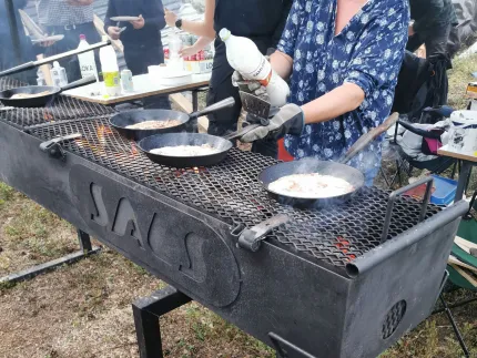 Person lagar mat i stekpannor på en stor kolgrill utomhus, med rök, eld och bord med saker i bakgrunden..