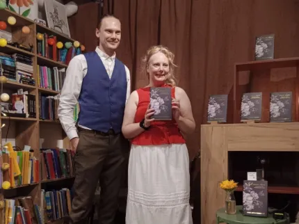 Två personer står framför en brun gardin och håller upp en bok, med bokhyllor och bokdisplay runtom..