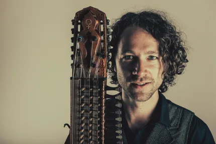 En man i mörkt lockigt hår iklädd mörk skjorta och väst, håller i en nyckelharpa.