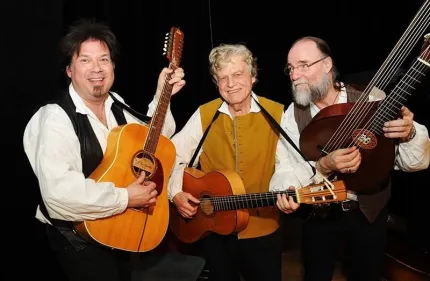 Tre musiker i västar och vita skjortor står på scen och spelar akustiska stränginstrument mot svart bakgrund..