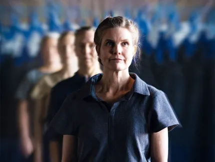 En kvinna med uppsatt hår, iklädd en blå kortärmad blus, bakom henne en man och två kvinnor på rad, i bakgrunden en grupp människor som är lite suddiga.