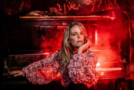 En kvinna i fluffig jacka sitter framför ett gammalt piano, omgiven av röd belysning och rök i mörkt rum..