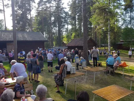 En stor grupp människor samlas på en gräsmatta mellan trähus och tallar, med bord och bänkar i solen..