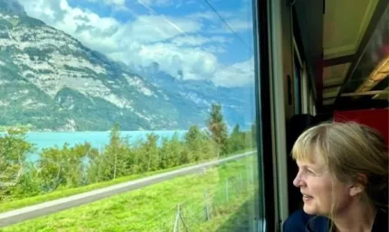 Vy genom tågfönster mot turkos sjö, gröna träd och berg under molnig himmel; passagerare syns till höger..