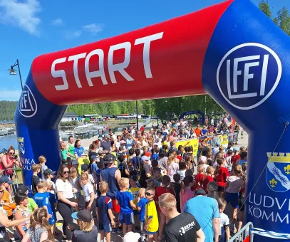 Stor uppblåsbar startbåge med texten START över en folkmassa av barn och vuxna vid ett lopp utomhus..