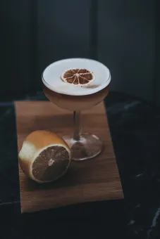 En skummig cocktail i coupeglas med torkad citrusskiva ovanpå, bredvid en halverad citron på träbricka..