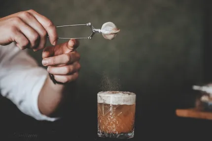 En hand håller en sil över ett glas cocktail medan fint pulver strös ned på skumtoppen mot mörk bakgrund..