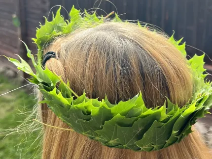 Närbild bakifrån av en person med blont hår som bär en grön lövkrans, med trädgård i bakgrunden..