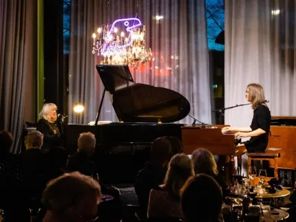 Två musiker spelar piano på en scen framför publik, med flygel, mikrofoner och en ljuskrona ovanför..
