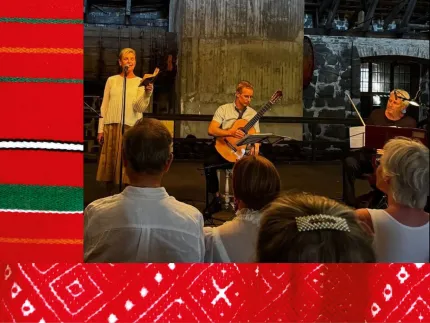 En kvinna som håller i en bok och står framför en mikrofon, en man som spelar gitarr och en man som spelar fiol, en folkdräktsdetalj i rött och grönt i bakgrunden.