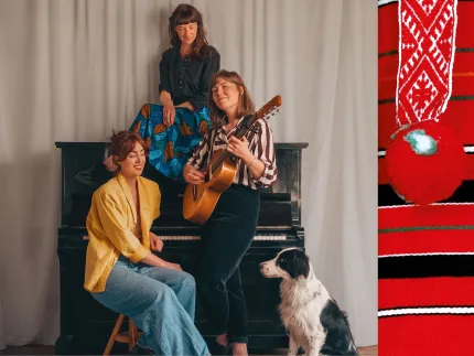 Tre kvinnor som sitter vid ett piano, en av dem håller i en gitarren sitter på pianot, en svartvit hund på golvet, en detalj från en folkdräkt i randigt svart, vitt rött och grönt samt ett band med en röd tofs.